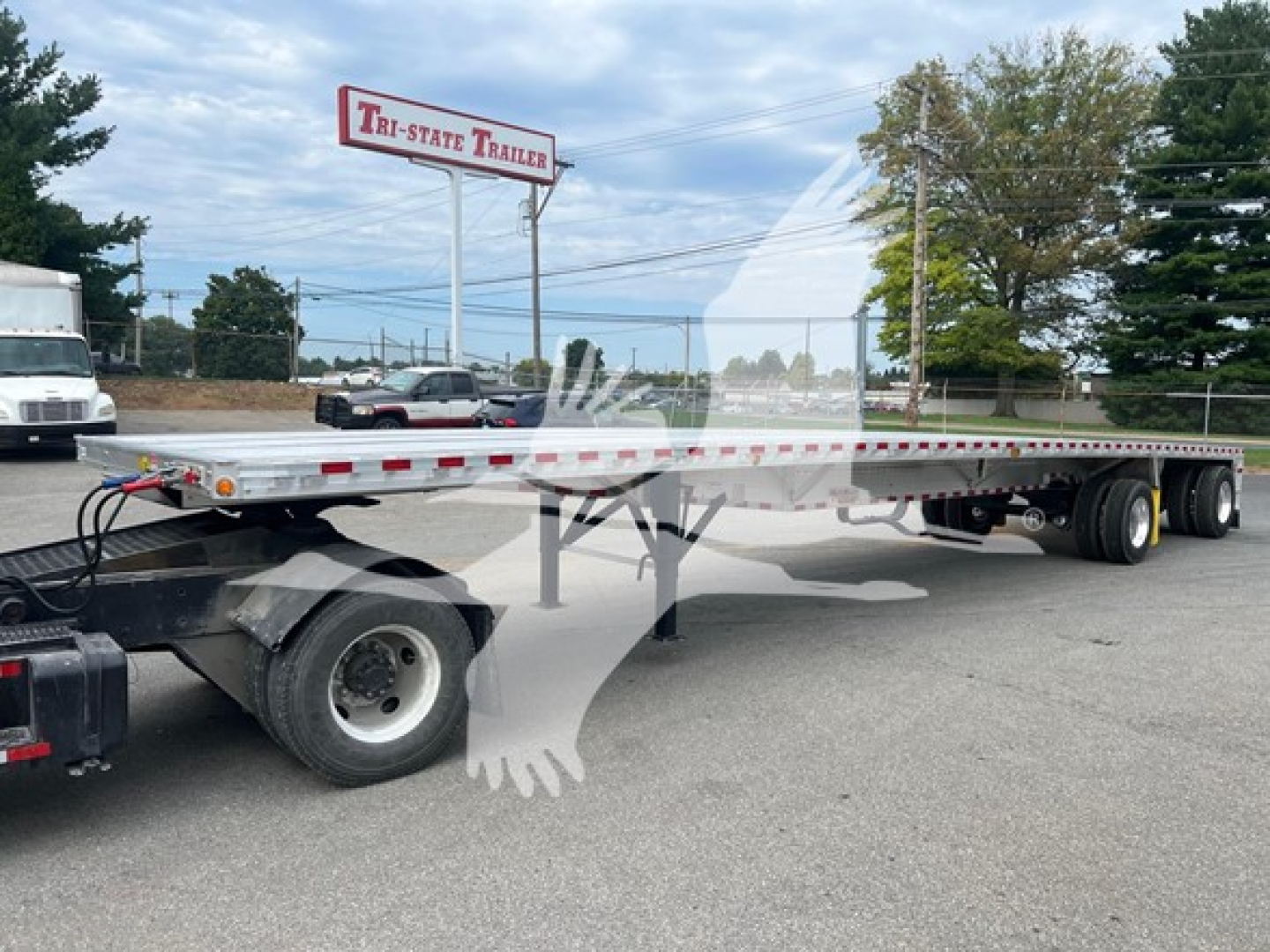 Flatbed trailer showing Gooseneck hitch and long aluminum deck with dual rear axles and tires, parked in lot near Tri-State Trailer sign.