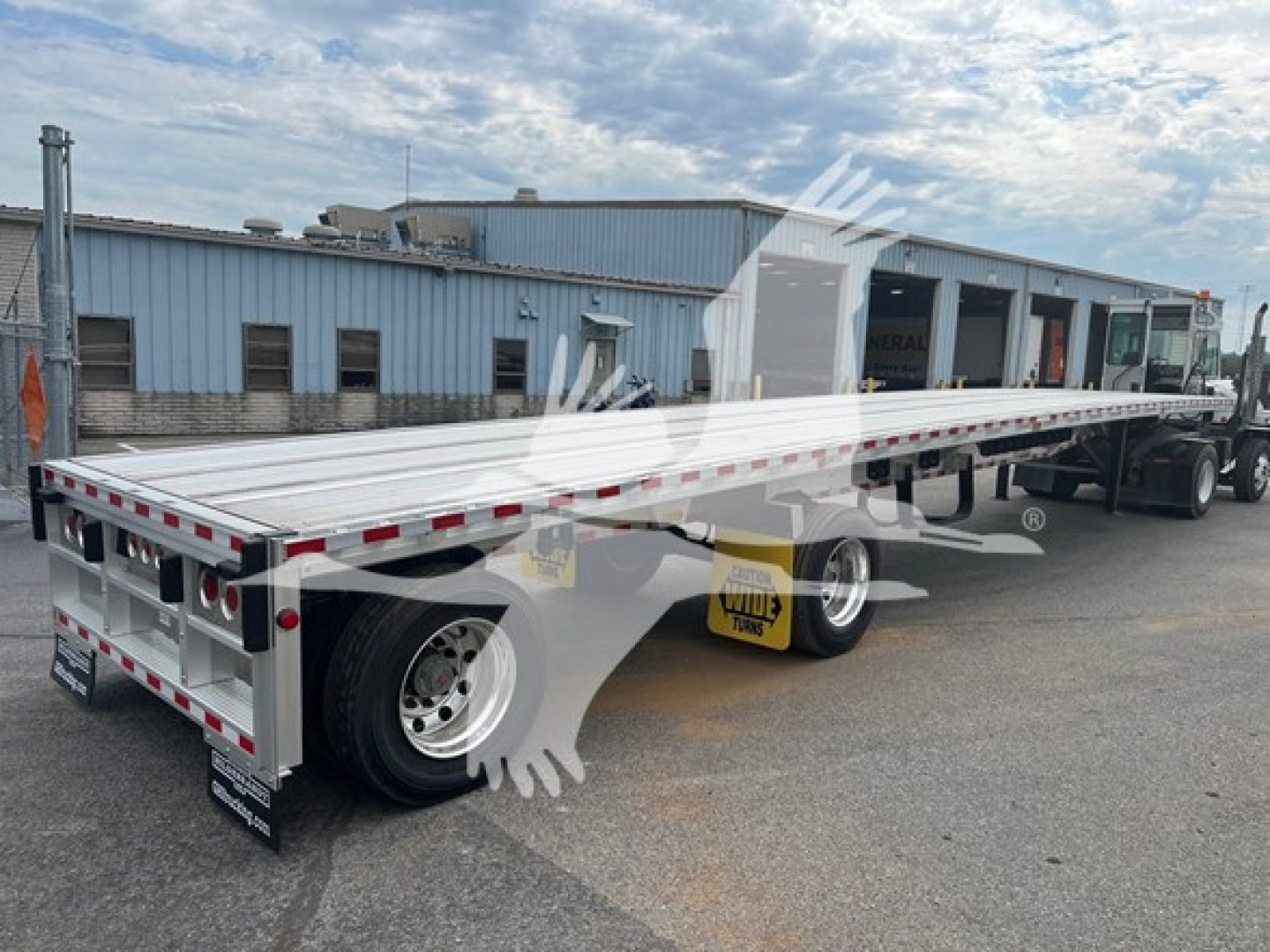 Maximise flatbed trailer showing long aluminum deck with red/white safety reflectors and rear underride guard, dual rear axles, empty hauler, warehouse in background.