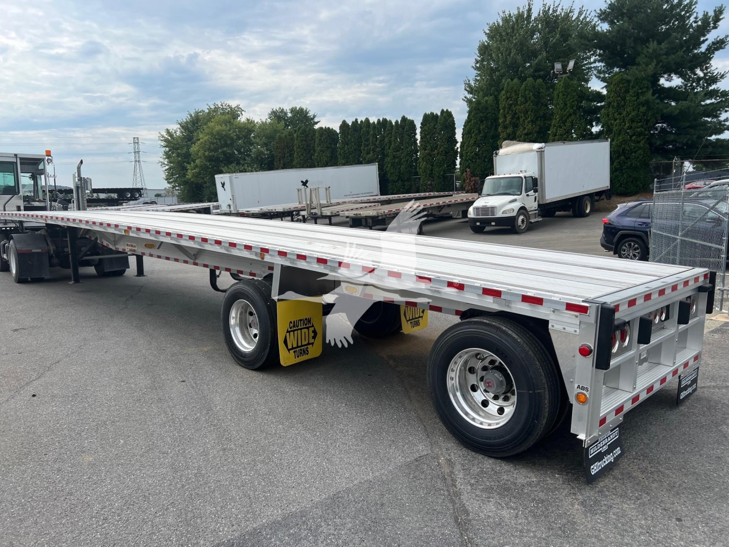 Flatbed trailer showing long aluminum deck with dual rear axles and wide tires, RGN-style with reflective tape and rear mud flaps.