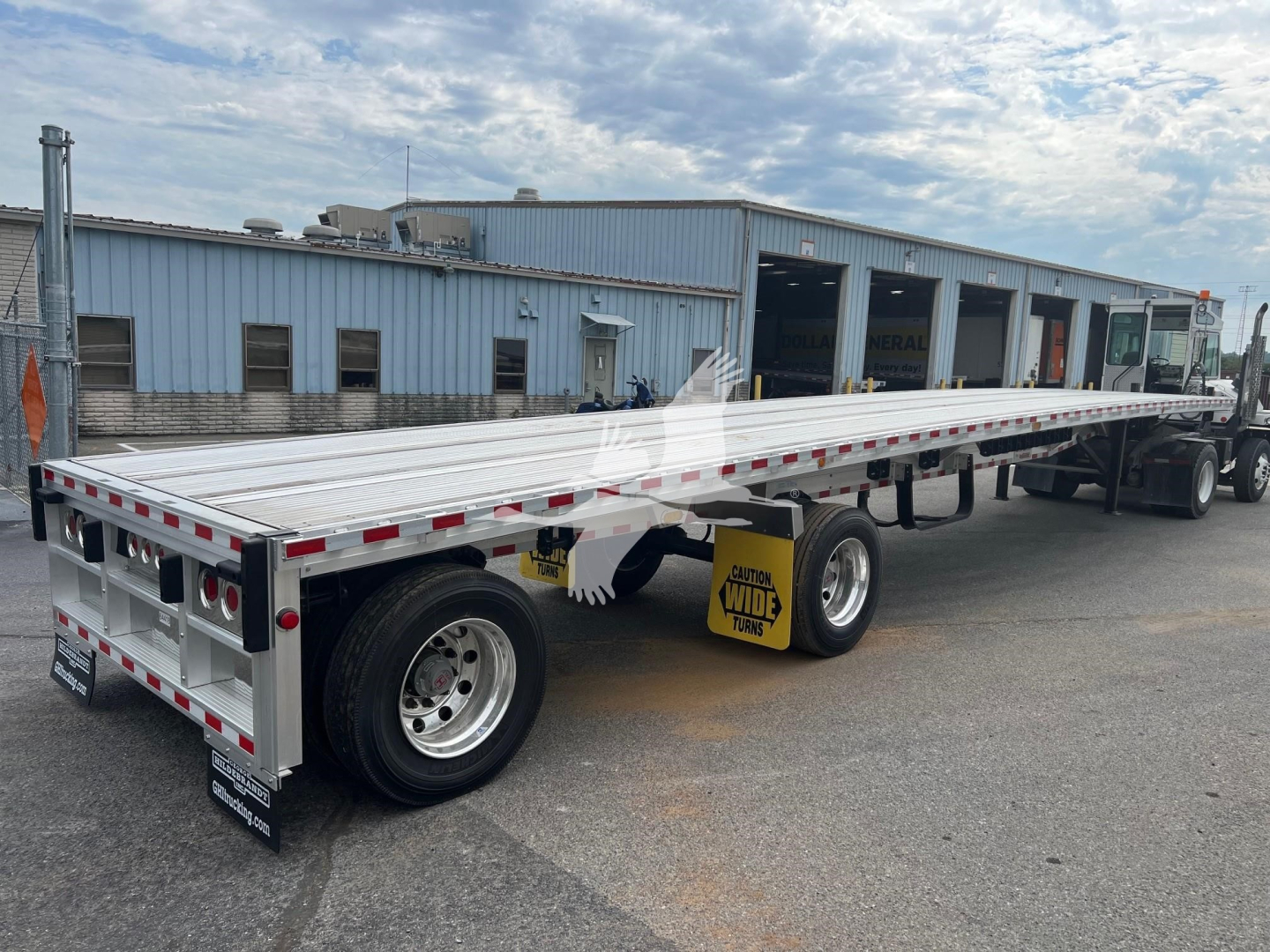 Flatbed trailer viewed from rear-left, long aluminum deck with reflective tape, dual rear axles, and yellow caution sign; truck cab visible in background.