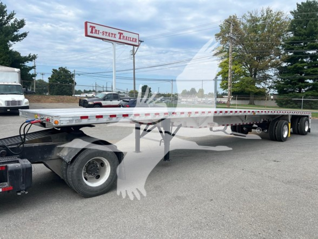 Flatbed trailer showing Gooseneck hitch and long aluminum deck with dual rear axles and tires, parked in lot near Tri-State Trailer sign.