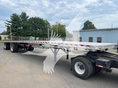 Flatbed trailer showing Maxmiser deck with dual rear axles and silver aluminum bed, black hitch area, and white wheels in a dealership lot