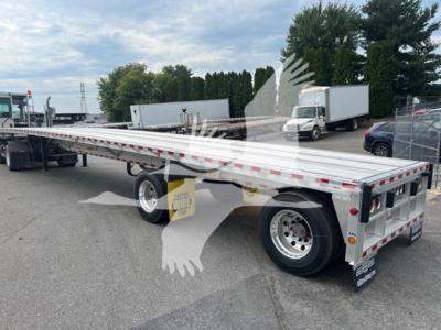 Flatbed trailer showing long aluminum deck with dual rear axles and e-track rails; white with red reflective safety tape; rear view shows bumper, taillights, and mud flaps; brand tag visible.