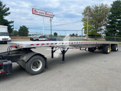 Flatbed trailer showing long aluminum deck with dual rear axles and undercarriage supports; Tri-State Trailer sign visible in background.