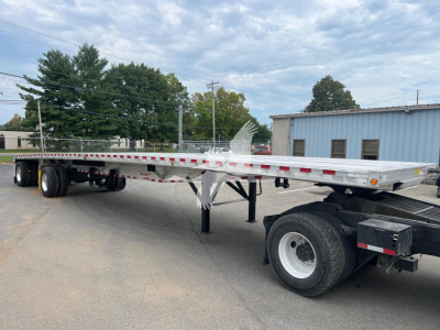 Flatbed trailer showing gooseneck hitch and dual rear axles with white wheels under a silver deck; side rails and reflective tape; clear blue sky in background.