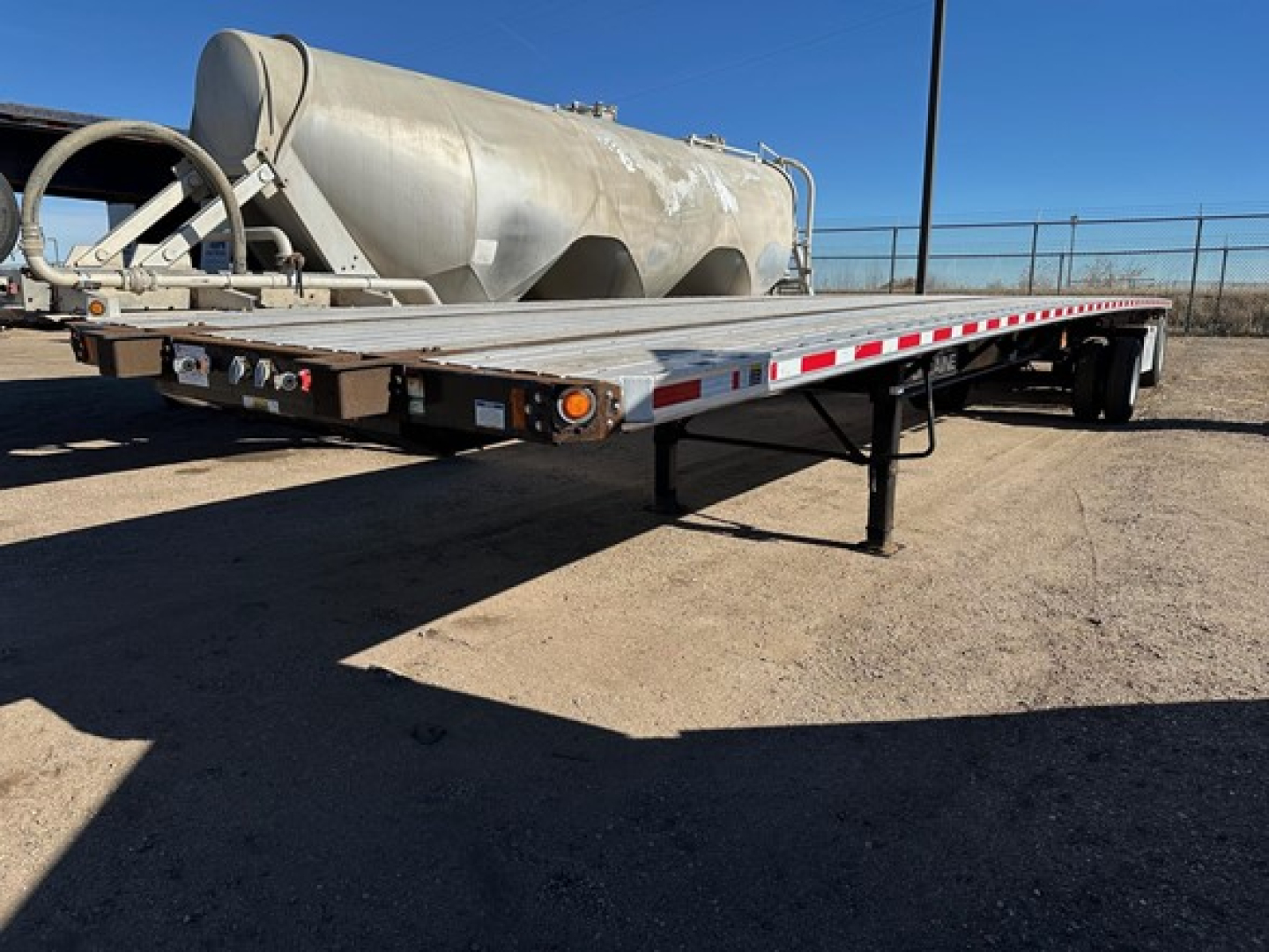 Flatbed trailer showing large cylindrical tank on the rear with a long flat deck, low angle view from left front, side markers and taillights visible.