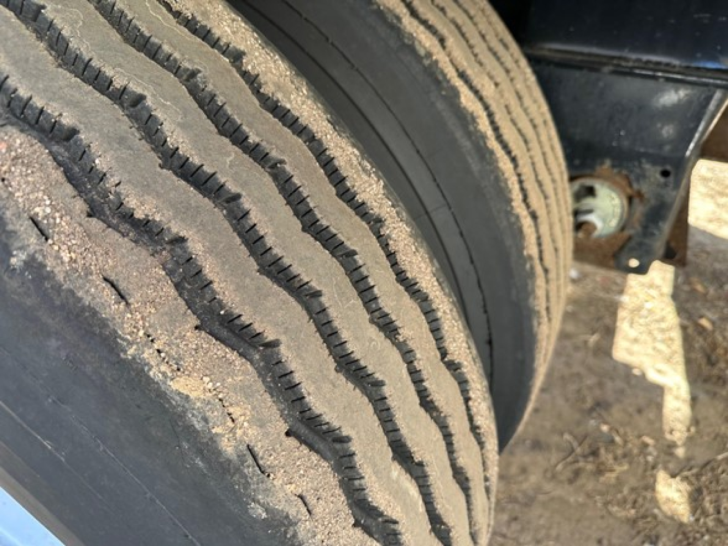 Trailer tire and wheel well close-up showing tread and dirt on the sidewall with chassis and mounting bracket in background.