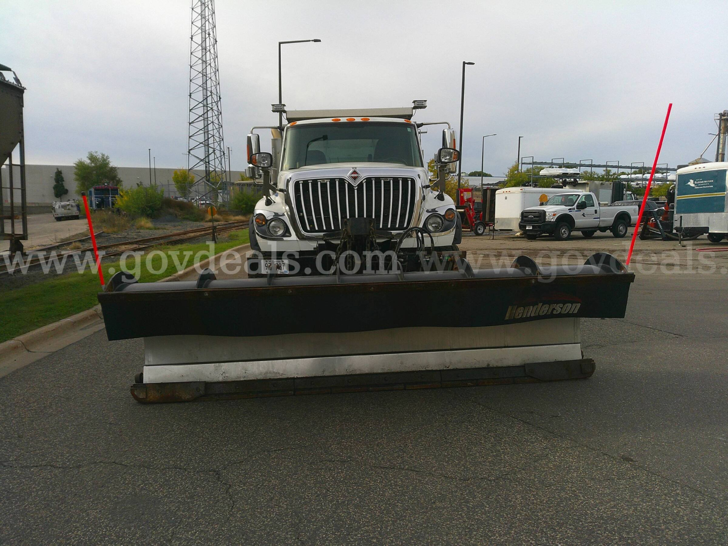 Truck with a large plow at the front; grille and headlights visible; plow shows the word 'Landsman' on the side and a white undercarriage beam; background includes other trucks and a parking lot.