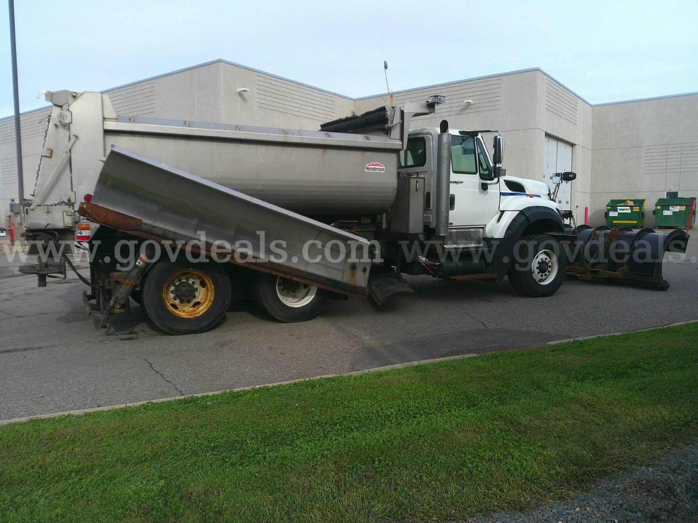 International WorkStar 7600 dump truck with white cab and gray dump body, plow/truck bed tilted, visible side label, in a lot near a building.