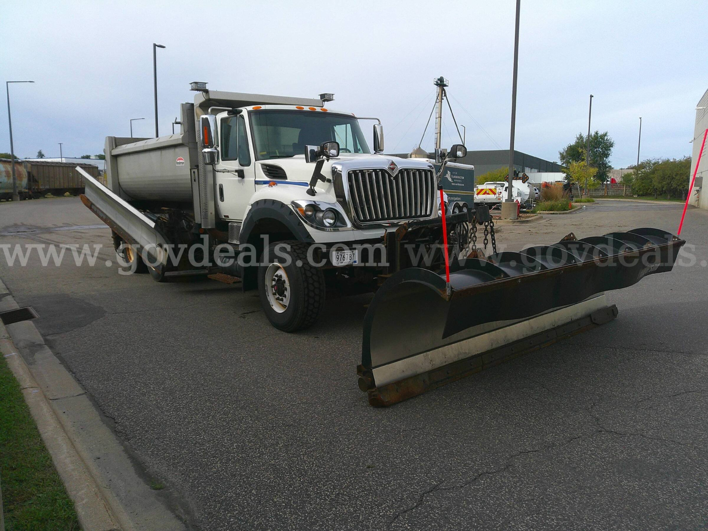 Truck with plow attachment showing International WorkStar 7600, front grille, dual rear wheels, large snow plow blade in foreground, utility bed on truck, overcast sky.