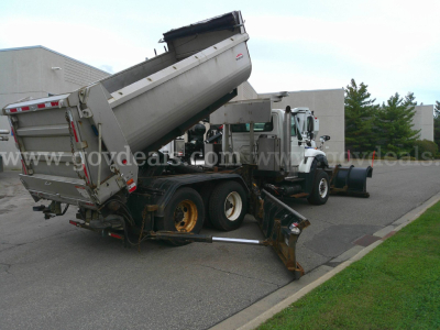International WorkStar 7600 dump truck with plow in a lot, rear dump bed raised, white cab, dual rear axles, side chute, and equipment around.