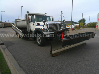 Truck with plow attachment showing International WorkStar 7600, front grille, dual rear wheels, large snow plow blade in foreground, utility bed on truck, overcast sky.