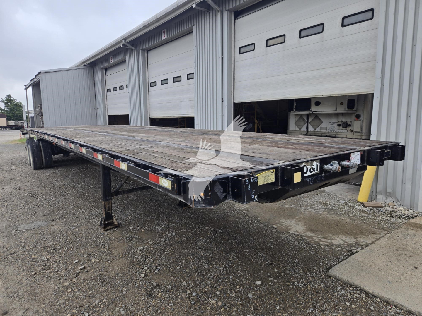 Flatbed trailer showing a long steel deck with dual rear axles and a hydraulic docking area near the cab; black frame with yellow corner post and reflective striping.