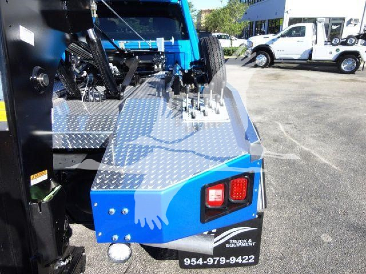 Ram 5500 SLT truck bed area with diamond plate platform and controls, rear taillight visible, blue body panel; winch equipment and nearby service lot in background.