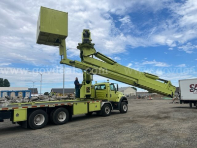 International 7600 bucket truck with extendable boom on a multi-axle trailer, green color, bucket elevated, clear sky.