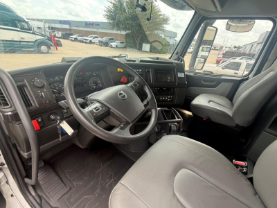 Volvo VNR truck cab interior showing steering wheel, dashboard, gauges, center console, and driver seat; visible exterior lot with other trucks through the windshield.