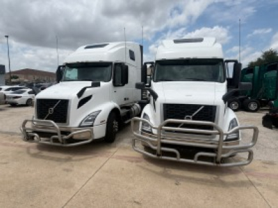 Two white Volvo VNR trucks with front guards parked side by side in a lot, cab-over design, standard sleeper configurations.