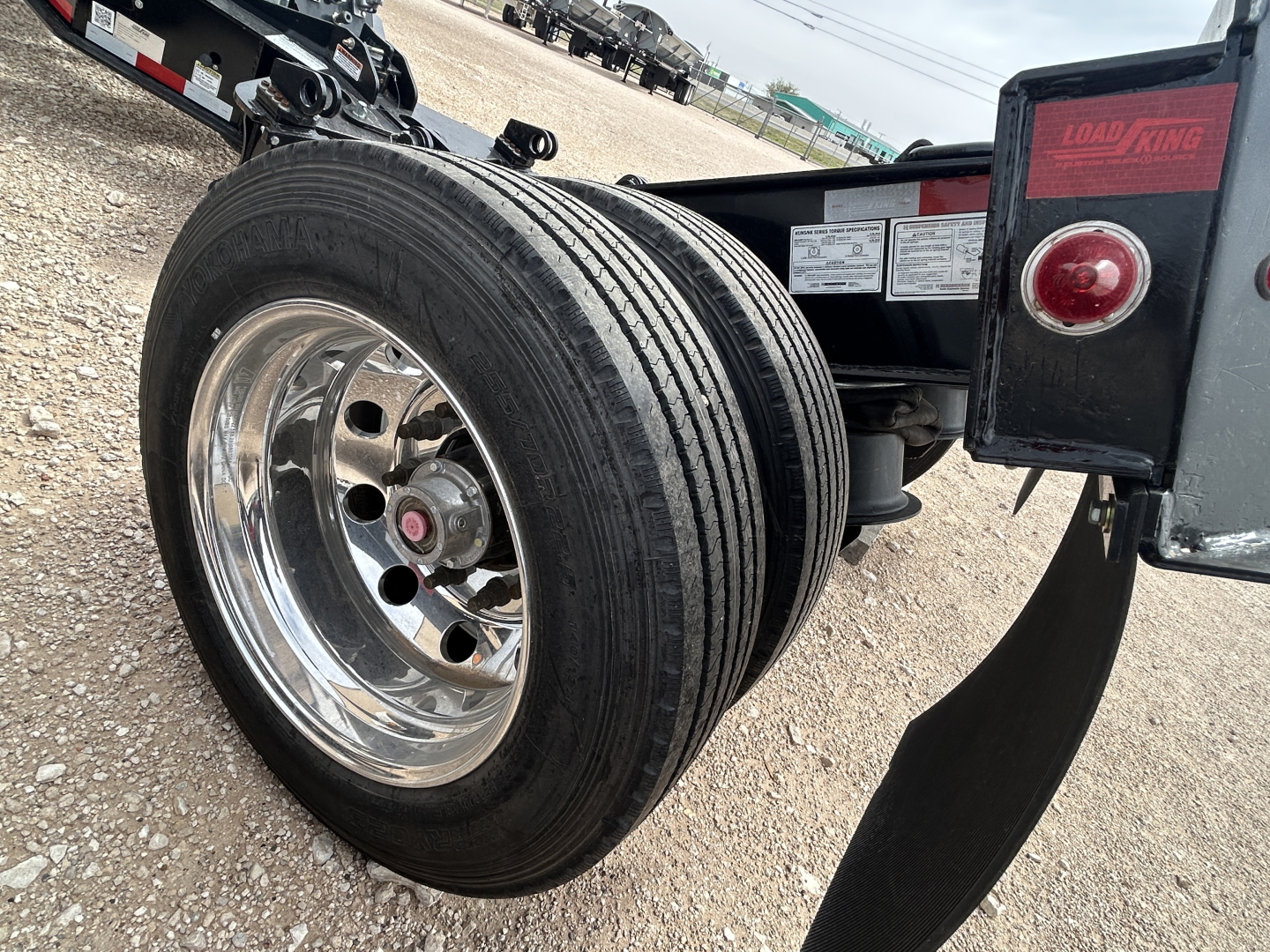 Load King trailer wheel and dual axle area showing a chrome wheel with black tire, fender, and Load King branding plate on the rear corner with a red taillight and safety decals.