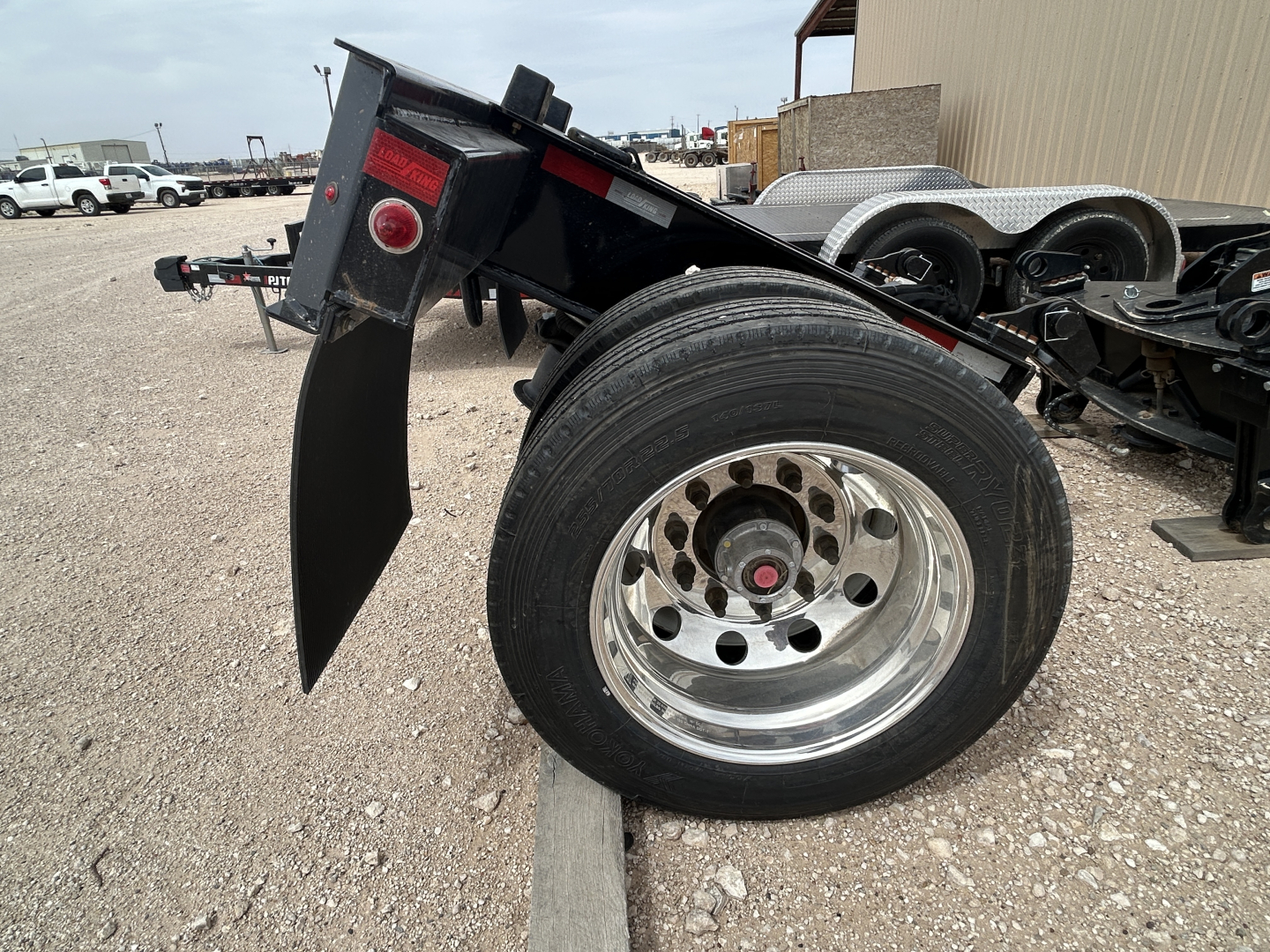 Load King trailer axle area showing a large rear tire and flip axle, chrome wheel, black frame, and rear fender showing part of the trailer in a gravel lot.