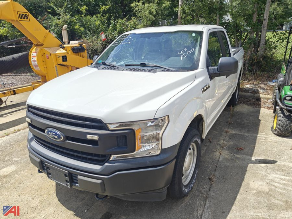 Ford F-150 pickup truck in white extended cab, front three-quarter view, blank plate, in a lot with a yellow crane and some trees in background.