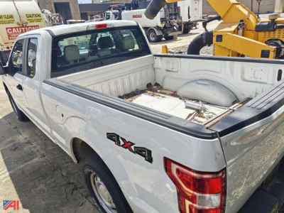 Ford F-150 pickup truck extended cab 4x4 with empty bed, white paint, visible tailgate and 4x4 badge, parked in lot with heavy equipment in background.