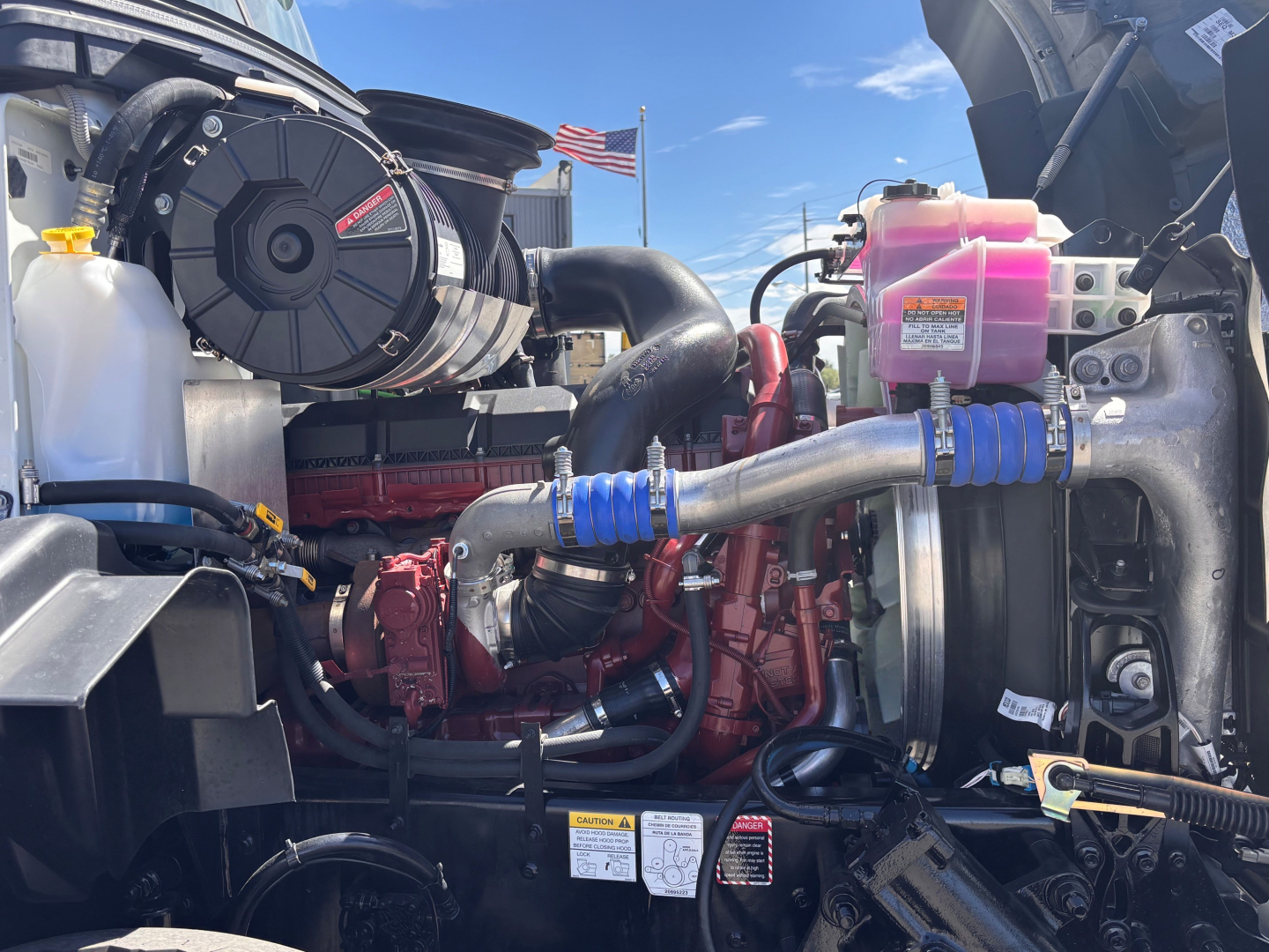 Engine bay of a 2027 Mack GR64B roll-off truck showing turbocharger, intercooler hoses, pink coolant reservoir, black air intake housing, red engine block, blue hose clamps, and various labels.