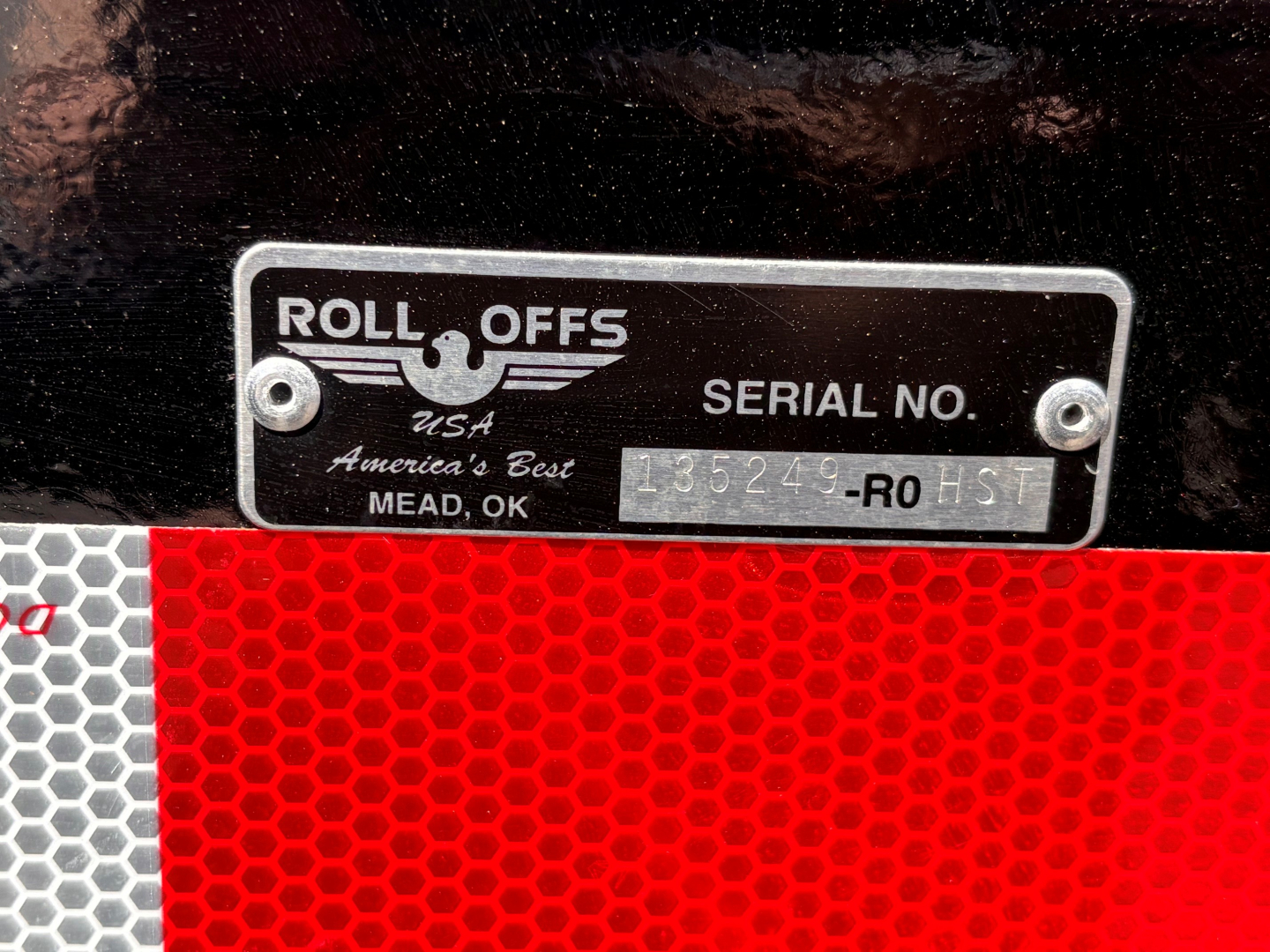 Roll-offs USA plate with serial number and MEAD, OK location; shows 'ROLL OFFS' logo and 'SERIAL NO.' field with characters; black plate mounted on equipment with red honeycomb reflector below.
