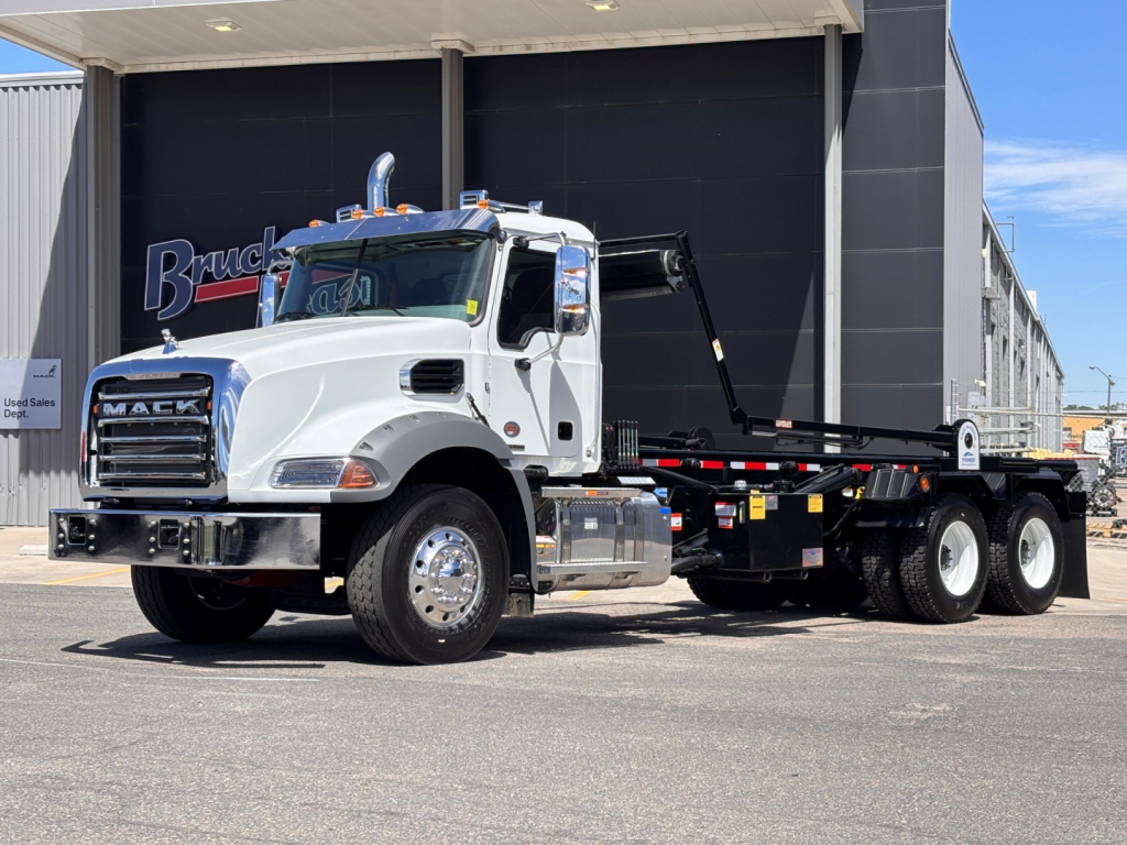 White 2027 Mack GR64B roll-off truck in front of a dealer building, with a black hook-and-flatbed body and chrome wheels.