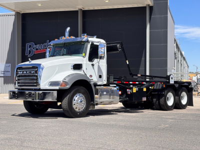 White 2027 Mack GR64B roll-off truck in front of a dealer building, with a black hook-and-flatbed body and chrome wheels.