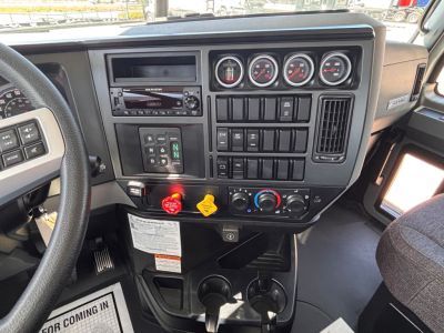 Dashboard of a 2027 Mack GR64B showing gauges, switches, and controls. Center panel has multiple round gauges, climate controls, and a digital readout; steering wheel left foreground.