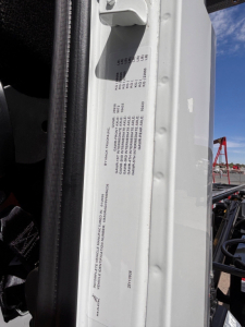 Label sticker on vehicle door frame with small printed text and numbers, part of a Mack GR64B roll-off truck showing VIN area and serials.