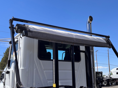 Truck cab front side showing chrome exhaust stack and rolloff hoist mechanism on a white 2027 Mack GR64B in-service cab area.