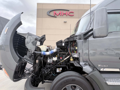 Kenworth T680 engine bay with hood open showing engine, intake piping, and exposed components; cab area beside front wheel; dealership MHС building in background.