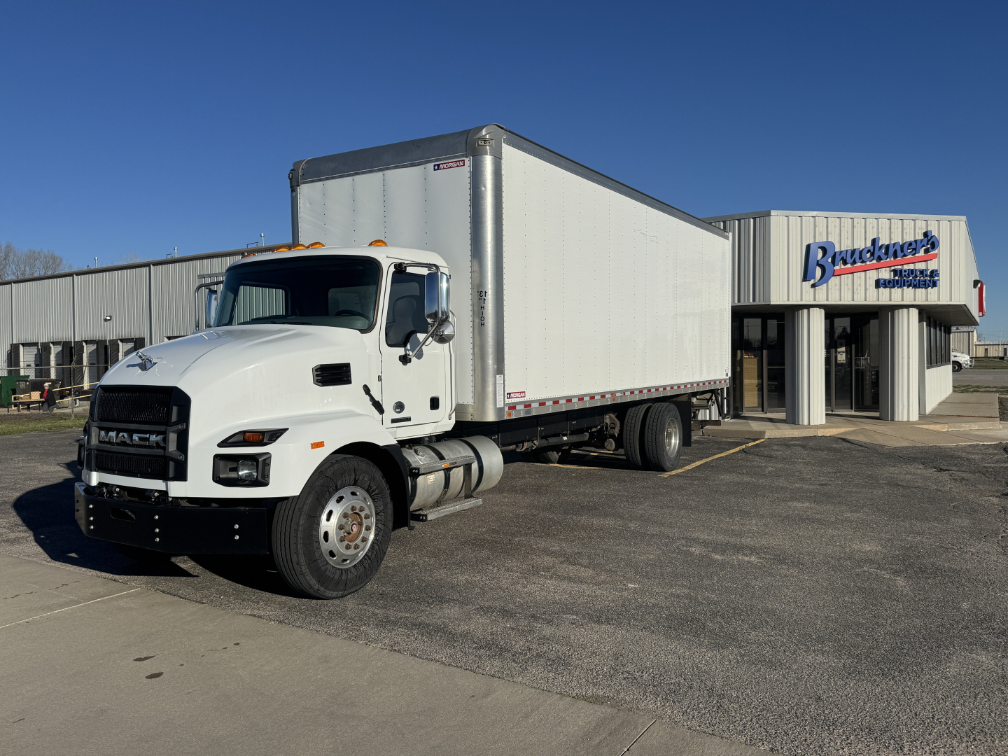 Truck with box trailer in dealership lot. Front showcases Mack MD7/Md6 cab, large white box trailer attached. Building in background with 'Brucker' sign.