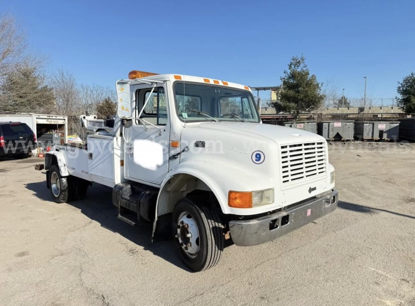 White International 4700 tow truck with flatbed, front right side view, VIN plate not visible, orange beacon lights on cab roof, large grille, round headlight assemblies, used condition.