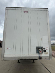 Rear view of a white Wabash trailer van showing a rear access door with latching mechanism and VIN? decals; compact corner panels and support legs visible.