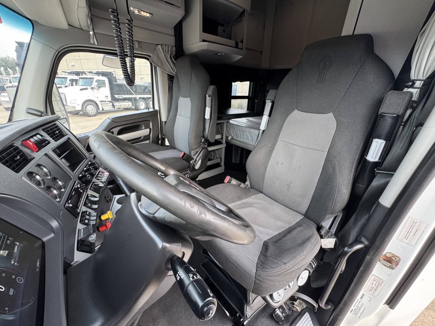 Cab interior of a Kenworth truck cab with steering wheel, driver's seat, and dashboard controls. Visible labels include truck interior panels and control buttons.