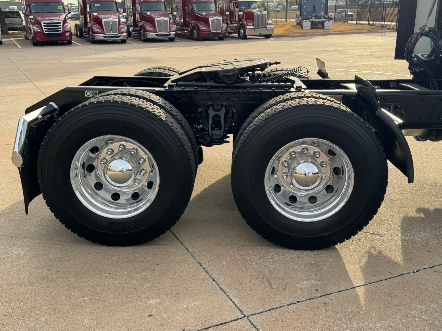 Kenworth T680 dual rear axle truck chassis with 22.5 inch wheels and chrome hubcaps, fuel lines, and frame rails in view at a dealership lot.