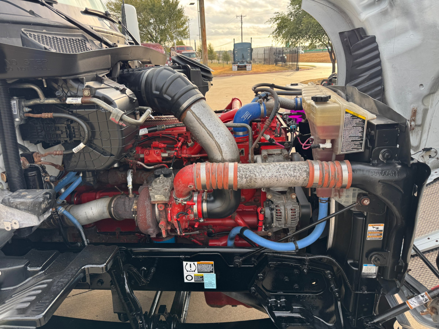 Kenworth T680 engine bay showing diesel engine with red intake manifold, blue and red hoses, black radiator frame, and various labels.