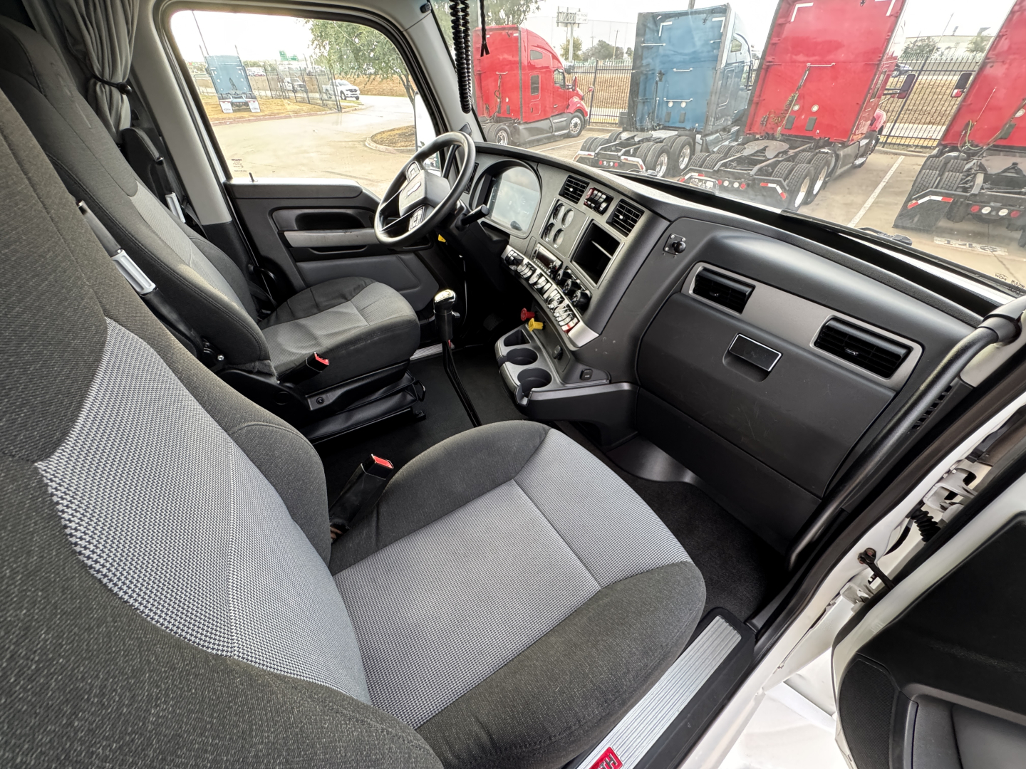 Interior of a 2022 Kenworth T680 cab showing driver seat, dash, steering wheel, and center console from the open door; gray fabric seats and dashboard with multiple controls.