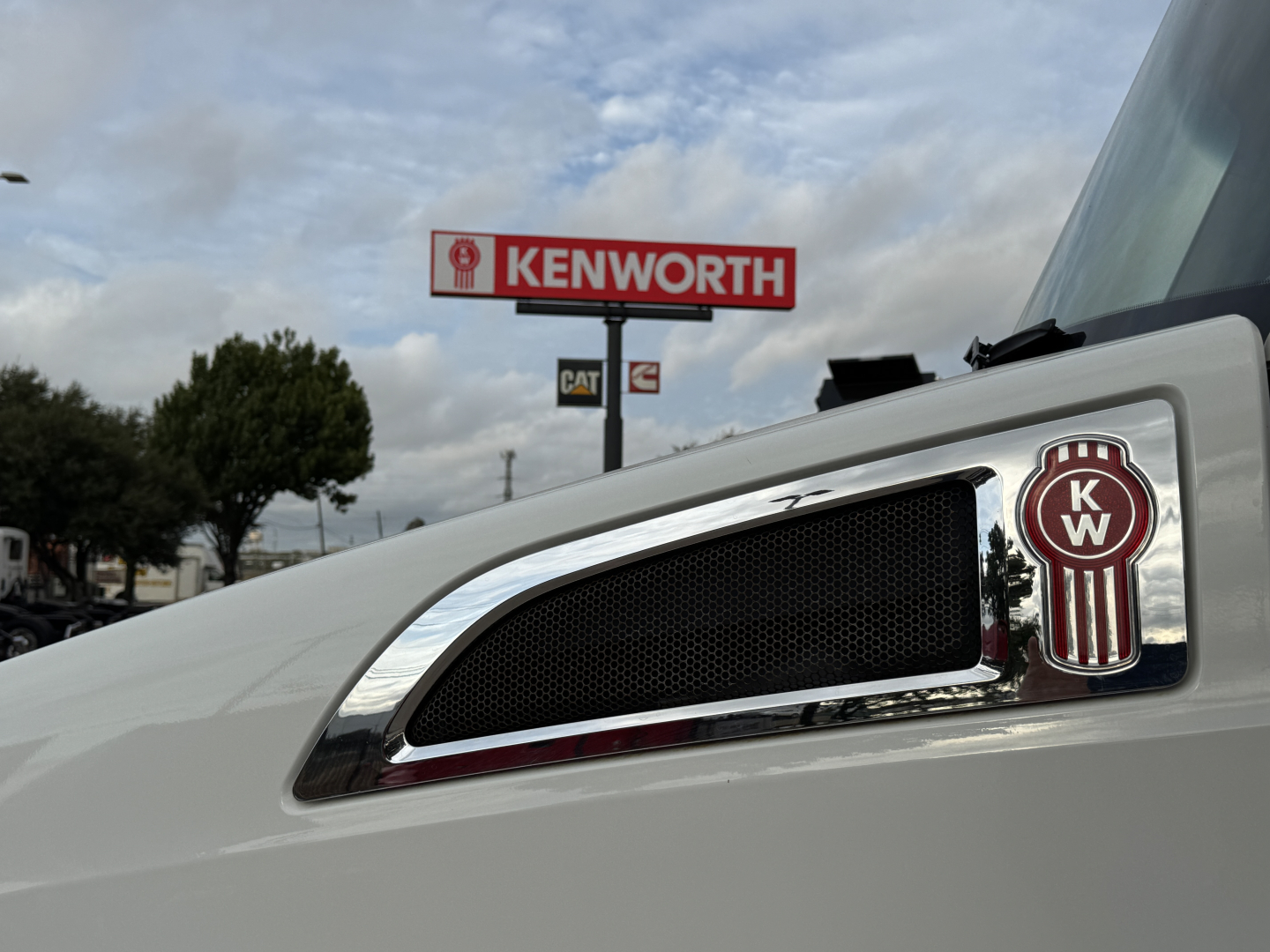 Kenworth logo badge on side of white 2022 Kenworth T680 truck with chrome grille accent and Kenworth sign in background.