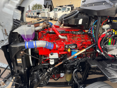 Kenworth T680 engine bay view showing red X15 engine with blue silicone hose, various fluid reservoirs, and wiring harnesses.
