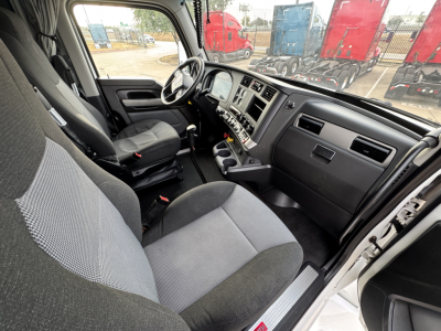 Interior of a 2022 Kenworth T680 cab showing driver seat, dash, steering wheel, and center console from the open door; gray fabric seats and dashboard with multiple controls.
