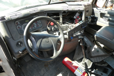Cab interior of a Freightliner Cascadia 113 showing steering wheel, dashboard gauges, exposed wiring, and center console with a red fire extinguisher nearby.