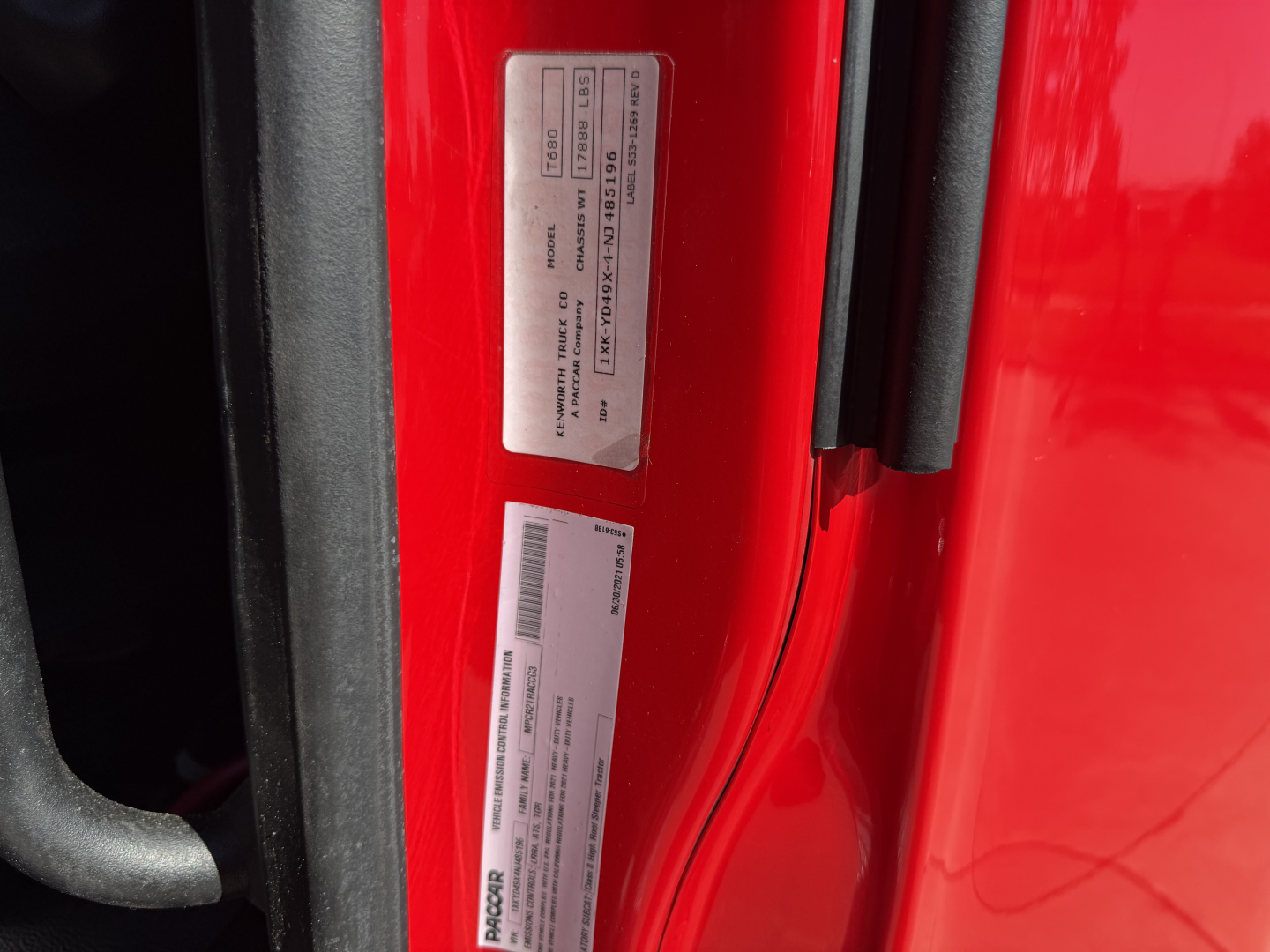 Dual vehicle labels on a red Kenworth T680 door jamb showing VIN and compliance stickers.