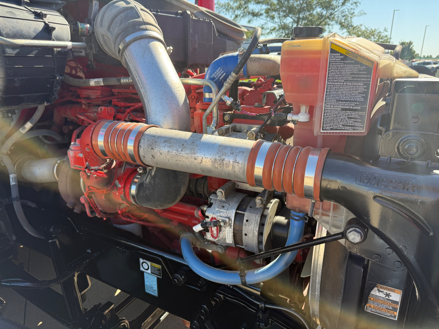 Kenworth T680 engine bay showing turbocharger and exposed hoses and hydraulic reservoir with warning label.