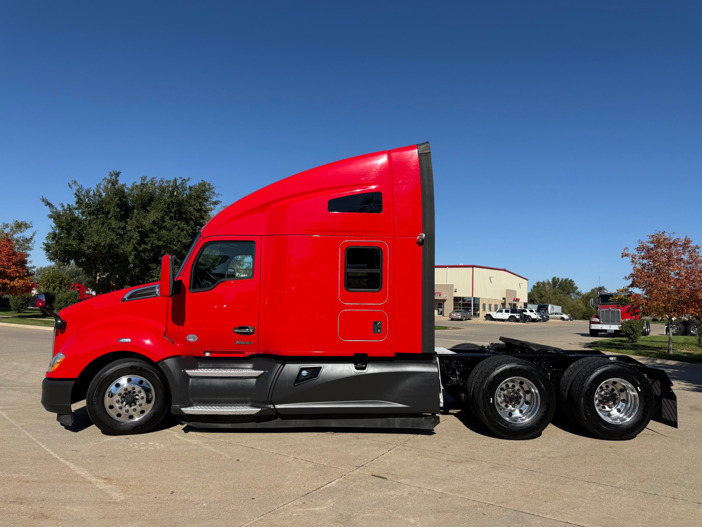 Kenworth T680 tractor unit in red side profile with sleeper cab, black lower panels, triple rear axles visible, chrome wheels.
