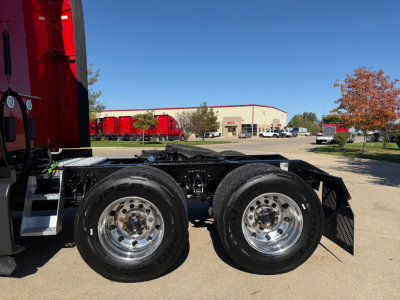 Kenworth T680 chassis with dual rear truck tires and wheel rims visible, cab extension on left, parking lot backdrop with trees and building in distance.