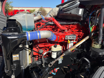 Kenworth T680 engine bay view showing Cummins X15 badge and various hoses, reservoirs, and wiring in red-painted engine area.