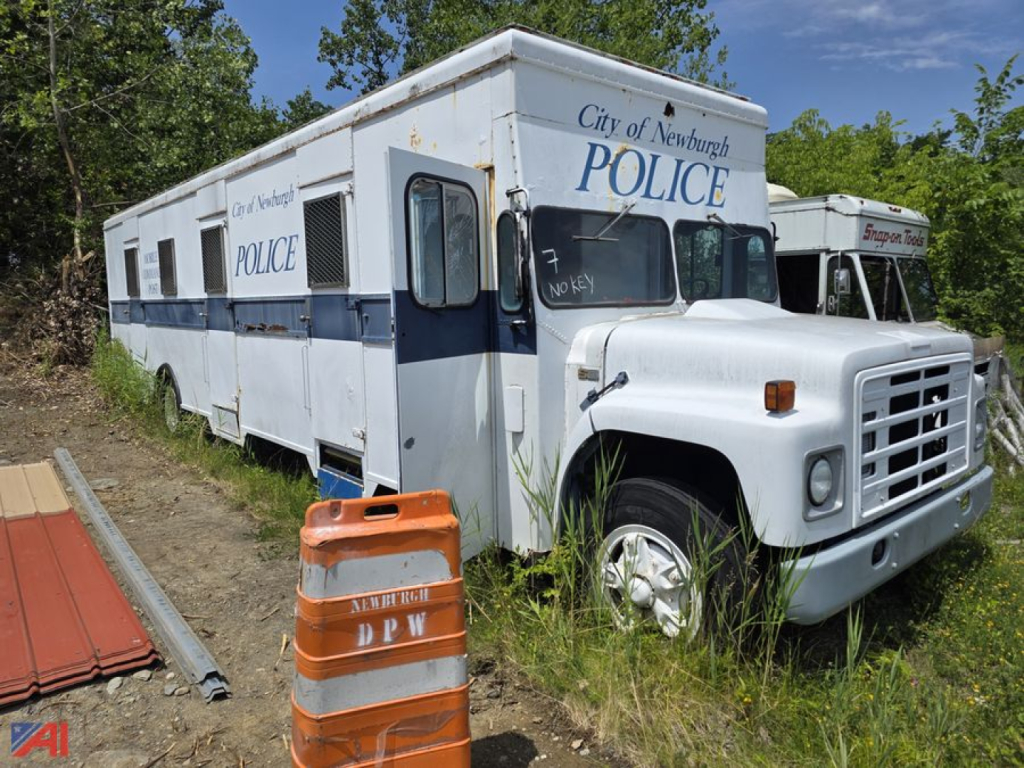 Mobile police station truck with City of Newburgh POLICE markings on white and blue exterior, side windows, and orange roadside barrier in foreground.